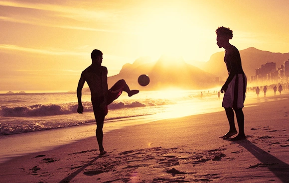 Silhueta de dois jovens jogando altinha na praia durante o pôr do sol, com o Morro Dois Irmãos ao fundo no Rio de Janeiro. Imagem em estilo documental e autêntico para ilustrar o marketing branding e a estratégia de marketing de conteúdo da campanha da Nike analisada pela Quel Chic.
